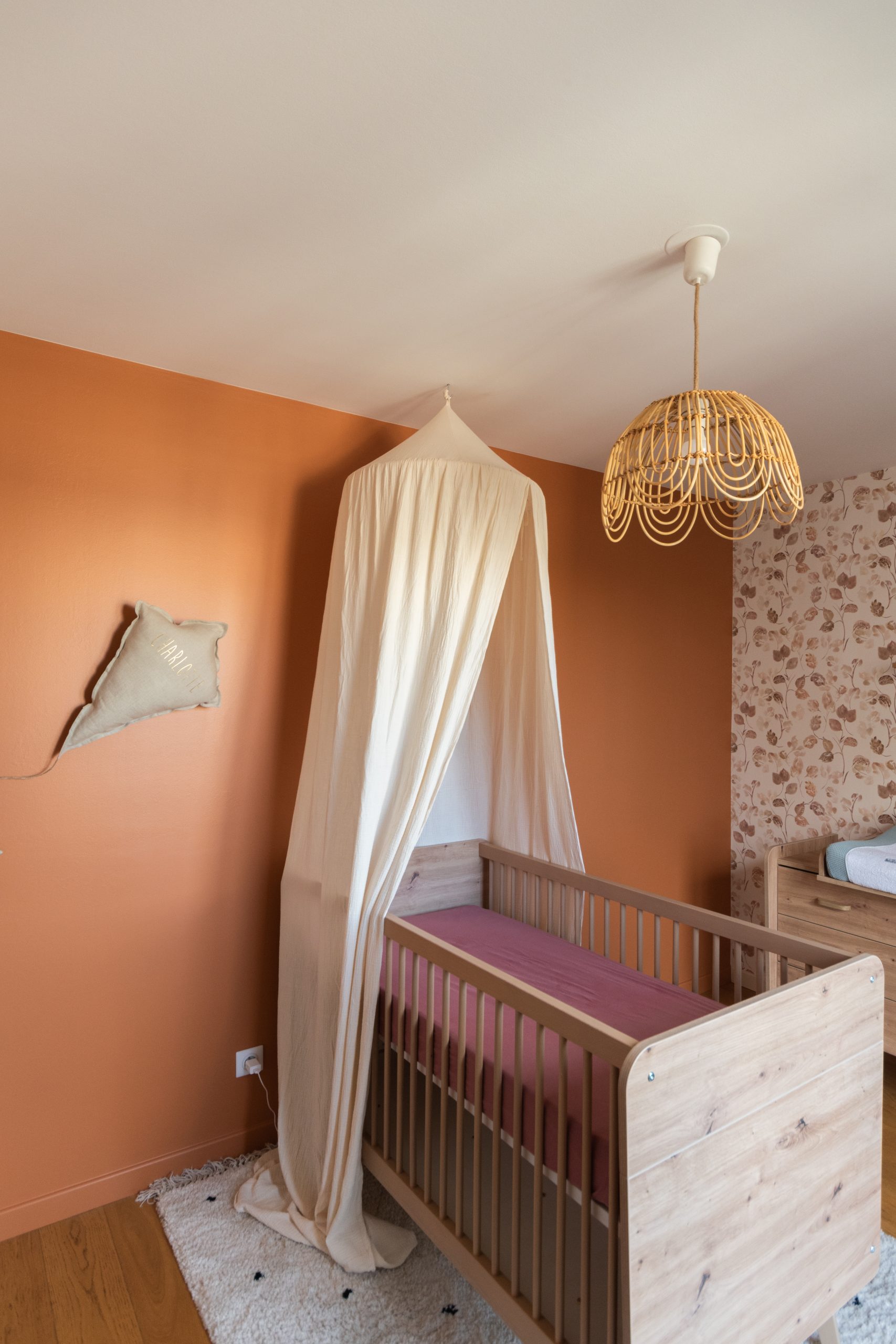 Photo du projet Reine qui montre le lit évolutif de cette chambre d’enfant mixte, le ciel de lit beige et la suspension en forme de fleur, en rotin. Le tout sur un fond de mur Terracotta.